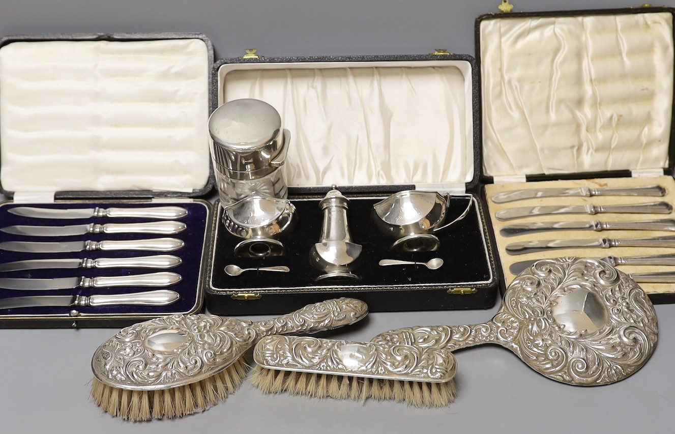 A cased 1950's silver three piece condiment set, two cased sets of six silver handled tea knives, a silver mounted glass salts bottle, silver mounted hand mirror and two brushes.